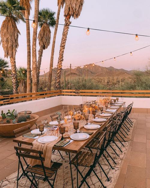 A beautifully set outdoor dining table with candles, surrounded by palm trees and mountains at sunset.