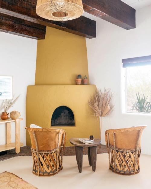 Cozy living room with a yellow fireplace, two woven chairs, a wooden table, and plants in a bright, airy space.