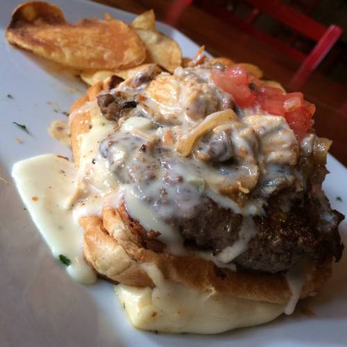 A hearty sandwich with a beef patty, melted cheese, topped with tomatoes and crispy onions, served with potato chips.
