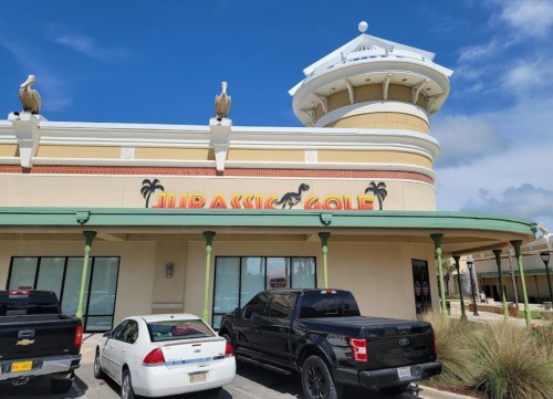 Exterior of a building with "Jurassic Golf" sign, featuring palm trees and a decorative tower, parked cars in front.