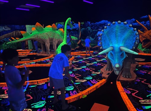 Children playing mini-golf in a vibrant, dinosaur-themed glow-in-the-dark setting.