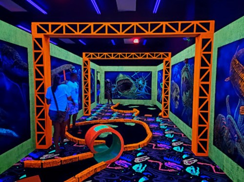 A vibrant glow-in-the-dark mini-golf course with colorful walls and a winding path, featuring ocean-themed decorations.