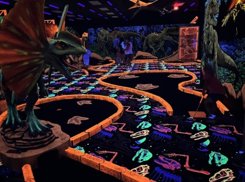 A glow-in-the-dark mini-golf course featuring dinosaur sculptures and vibrant, colorful designs on the floor.