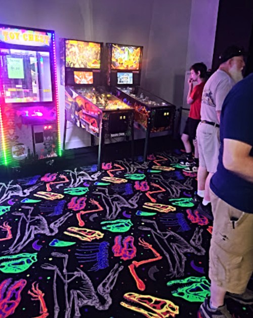 A brightly lit arcade with pinball machines and a toy crane game, featuring a colorful, patterned floor.