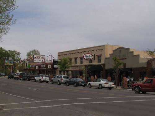 A street view featuring shops and parked cars along a small-town main street under a cloudy sky.