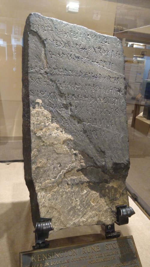 A large stone tablet with ancient inscriptions displayed in a glass case, labeled "Kensington Rune Stone."