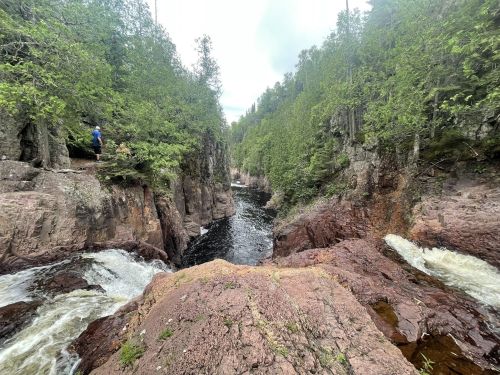 A scenic view of a river flowing through rocky cliffs, surrounded by lush greenery and a person standing nearby.