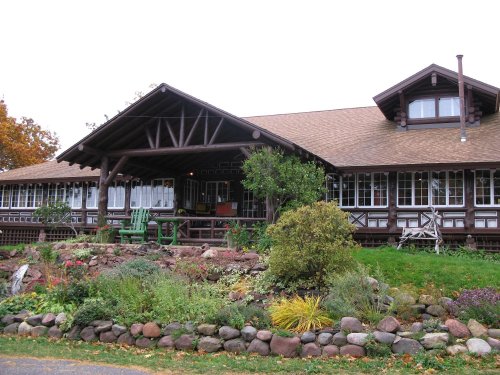 A large wooden lodge with a sloped roof, surrounded by gardens and stone landscaping, under a cloudy sky.