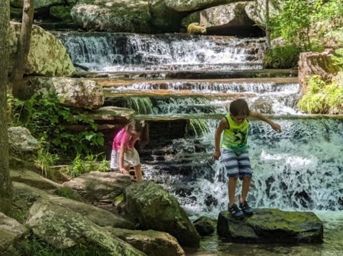 Two children play near a cascading waterfall, exploring the rocks and water in a lush, green environment.