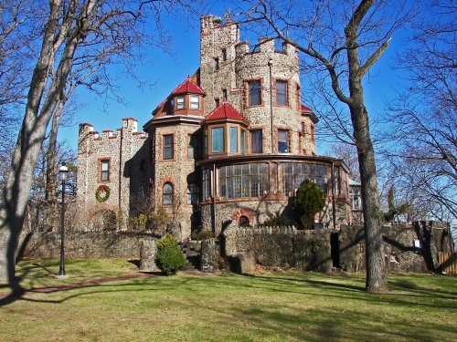 A stone castle with turrets and a red roof, surrounded by trees and a grassy lawn under a clear blue sky.