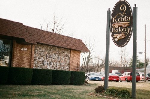 Exterior of Kruta Bakery featuring a stone wall and a sign, with parked cars in the background on a cloudy day.