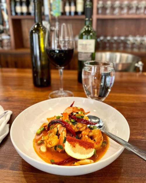 A bowl of shrimp dish with herbs and spices, accompanied by a glass of red wine and water, set on a wooden table.