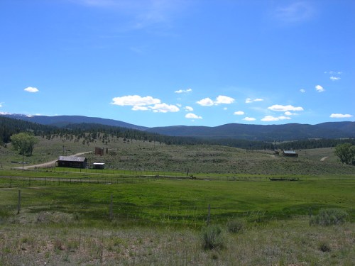 A scenic landscape featuring rolling green hills, a farmhouse, and distant mountains under a clear blue sky.
