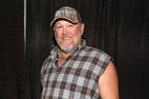 A smiling man in a plaid sleeveless shirt and camouflage cap stands against a black backdrop.