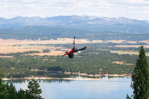 A person zip-lining over a scenic landscape with mountains, trees, and a lake below.