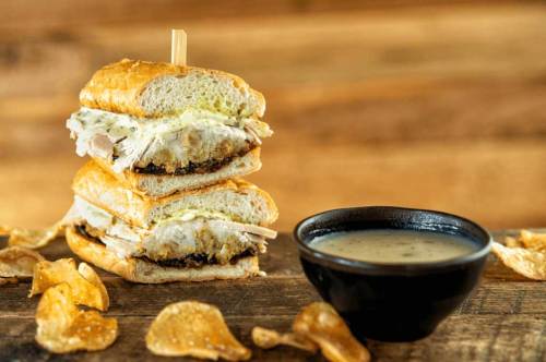 A stacked sandwich with turkey and creamy sauce, served with potato chips and a small bowl of gravy on a wooden surface.