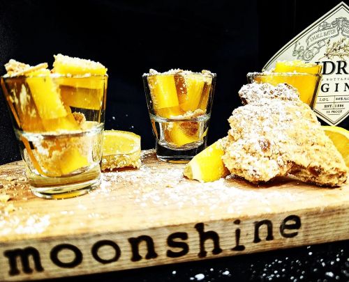 Three shot glasses filled with yellow moonshine and lemon slices, accompanied by a piece of ginger on a wooden board.