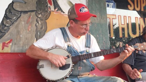 A man in overalls and a red cap plays the banjo on a porch, with a colorful mural in the background.