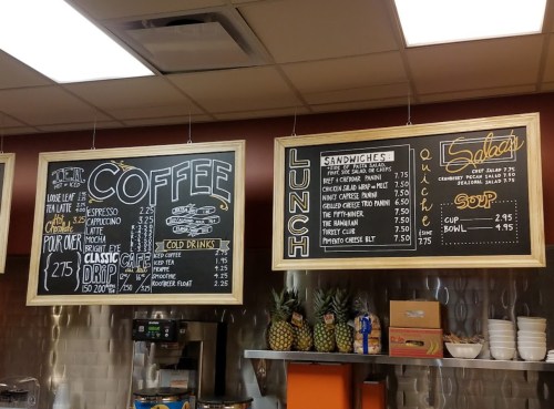 Menu boards displaying coffee, lunch, sandwiches, salads, and drinks with prices in a café setting.