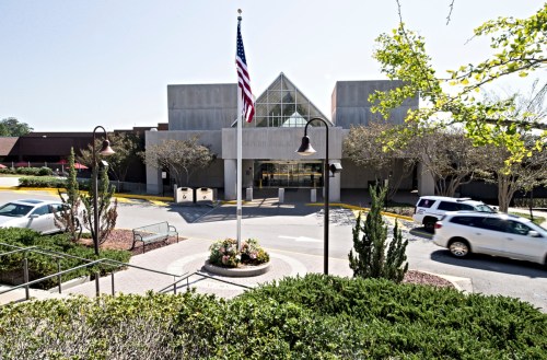 A modern building with a glass entrance, surrounded by trees and a flagpole, with a car passing by.