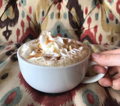 A hand holds a white cup of coffee topped with whipped cream and a sprinkle of cinnamon, set against a colorful patterned background.