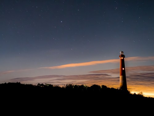 A lighthouse stands against a starry night sky, with soft clouds and a glowing horizon in the background.