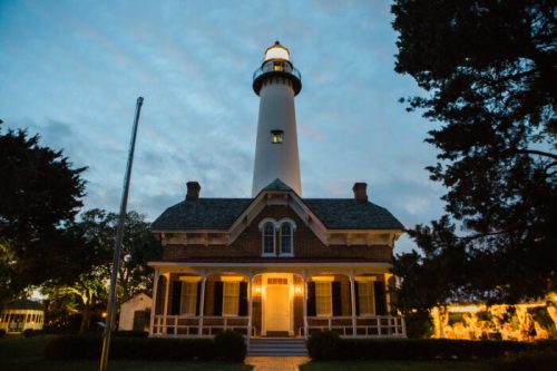 A historic lighthouse with a tall tower, illuminated at dusk, beside a charming brick house surrounded by trees.