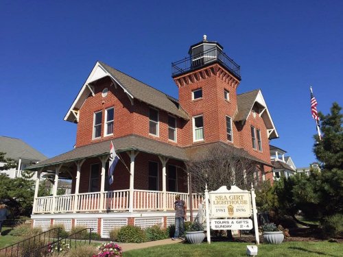 A red brick lighthouse with a white porch, surrounded by greenery and a clear blue sky, featuring a sign for tours.