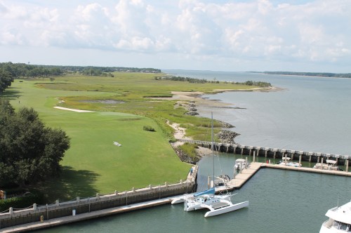 A serene coastal view featuring a marina, grassy shore, and calm waters under a partly cloudy sky.
