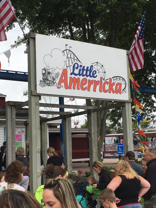 Sign for "Little Amerricka" amusement park with people gathered below, flags and rides visible in the background.
