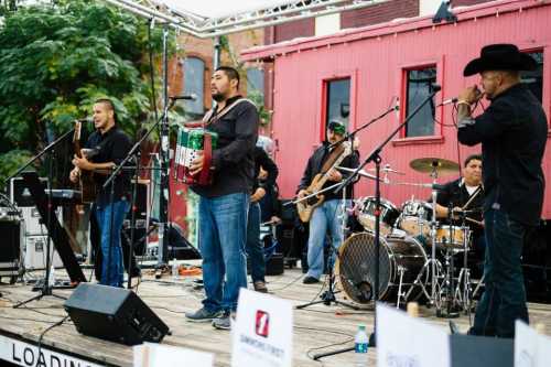 A lively band performs on stage with guitars, drums, and an accordion, set against a colorful backdrop.