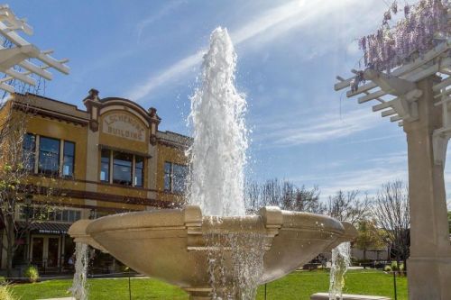 A stone fountain sprays water in a park, with a historic building and clear blue sky in the background.