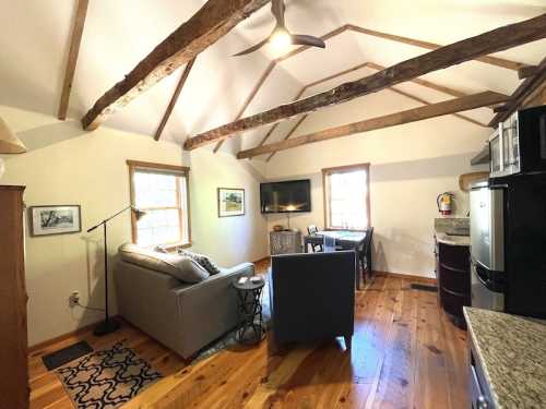 Cozy living space with wooden beams, a sofa, TV, and dining area, featuring warm wood floors and natural light.