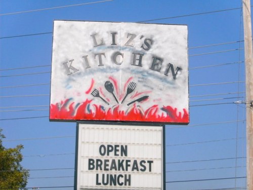 Sign for "Liz's Kitchen" with utensils graphic, advertising open for breakfast and lunch. Bright blue sky in the background.