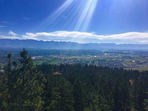 A panoramic view of a valley surrounded by mountains, with sun rays shining over a forested area.