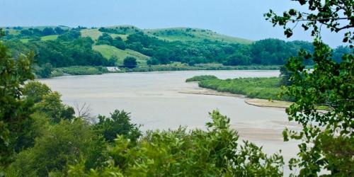 A serene river flows through lush greenery, with rolling hills in the background under a cloudy sky.