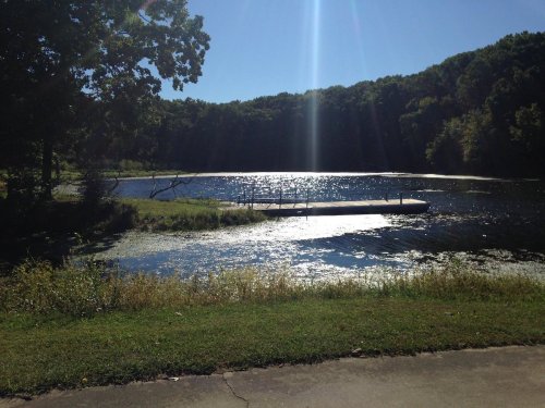 A serene lake surrounded by trees, with sunlight reflecting on the water and a small dock in the foreground.