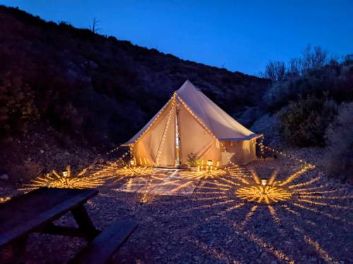 A beautifully lit tent surrounded by glowing lights, set against a twilight backdrop of hills and trees.