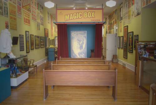 Interior of a theater called "Magic Box," featuring wooden benches, a stage with curtains, and vintage posters on the walls.