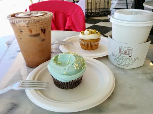 A table with two cupcakes, one blue with sprinkles and one white with pistachios, alongside iced coffee and a hot drink.