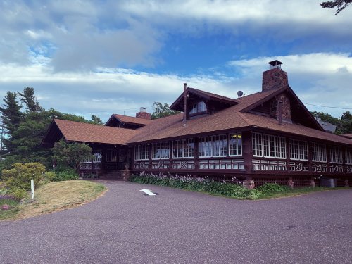 A large, rustic wooden lodge with a sloped roof, surrounded by greenery and a cloudy sky.