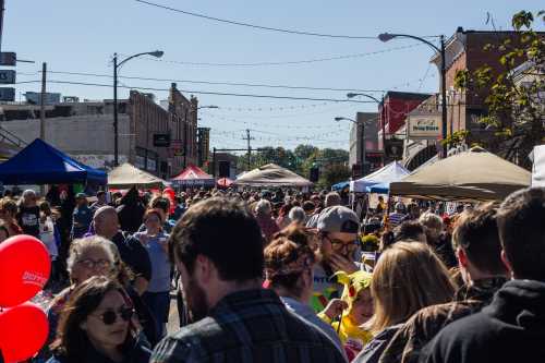 A bustling street fair with crowds, tents, and colorful balloons under a clear blue sky.