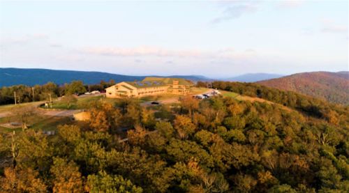 Aerial view of a building on a hill surrounded by colorful autumn trees and distant mountains under a clear sky.