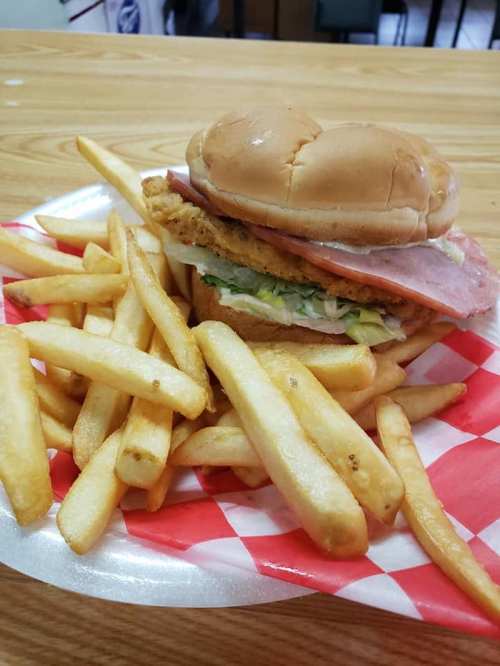 A sandwich with fried chicken, ham, lettuce, and a bun, served with a side of golden French fries on a red-checkered plate.