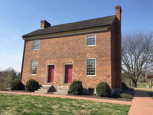 A two-story brick house with red doors, surrounded by grass and trees under a clear blue sky.