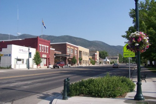A quiet small town street with shops, mountains in the background, and colorful flower baskets along the sidewalk.