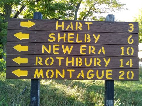 Wooden directional sign with arrows pointing left, listing distances to Hart (3), Shelby (6), New Era (10), Rothbury (14), and Montague (20).