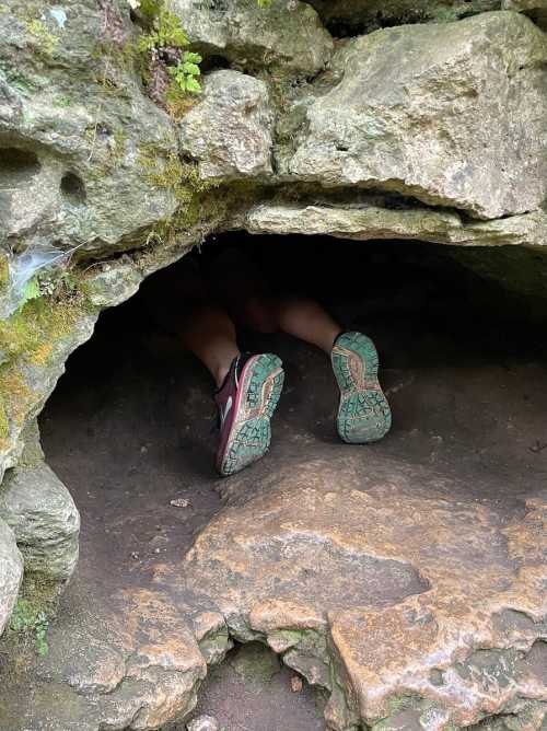 A pair of feet in hiking shoes sticking out from a small cave entrance in a rocky landscape.