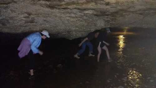 Three people wade through shallow water in a dimly lit cave, with rocky walls and a low ceiling above them.