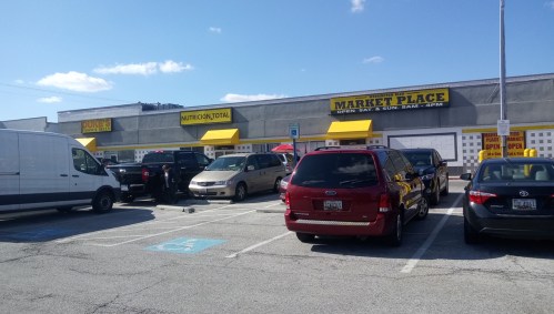 A parking lot filled with cars in front of a marketplace with yellow awnings and signage.